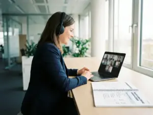 Fachkraft mit Kopfhörern im Video-Call am Laptop, daneben aufgeschlagenes Kurs-Workbook auf modernem Büroschreibtisch.